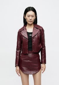 Vrouw draagt een donkerrode leren cropped jas en bijpassende minirok, gecombineerd met een zwart topje en hangende oorbellen, staand tegen een witte achtergrond.