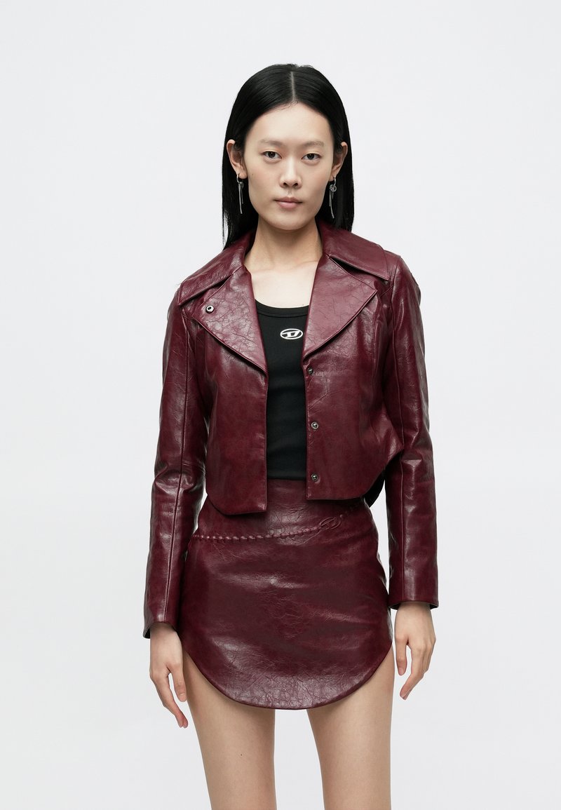 Mujer con chaqueta corta de cuero rojo oscuro y minifalda a juego, combinadas con un top negro y pendientes colgantes, posando frente a un fondo blanco.