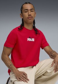 Rød bomulds t-shirt med et hvidt PUMA-logo, korte ærmer og rund halsudskæring, parret med lyse farvede bukser.