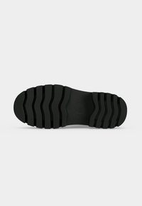 Semelle de chaussure noire avec de profondes rainures horizontales pour la traction, montrée à plat sur un fond blanc.