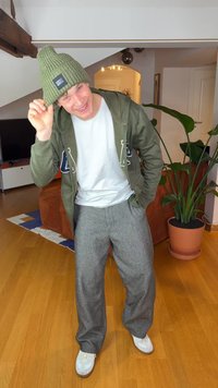 Grön stickad mössa, olivfärgad huvtröja med logotyp, vit t-shirt, grå mönstrade byxor och vita sneakers. Inomhusmiljö med en växt.
