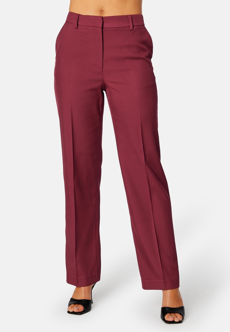 Femme portant un pantalon bordeaux taille haute, coupe droite, associé à des talons noirs ouverts à l'avant, sur un fond uni.