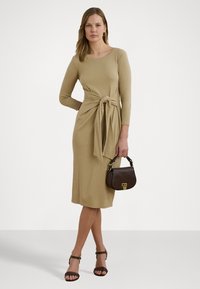 Robe tricotée beige à manches longues et taille cintrée par un lien. Associée à un sac à main marron et des sandales à talons sombres. Design longueur sol.