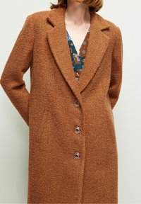 Femme portant un manteau marron texturé avec de grands revers et trois boutons visibles sur une robe à motifs bleus et bruns, les mains derrière le dos.