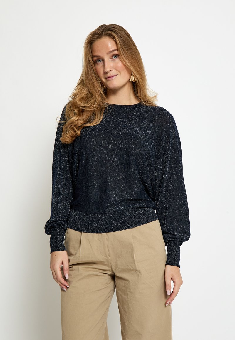 Maglione blu navy con trama metallica, vestibilità comoda, spalle scese e polsini a costine. Abbinato a pantaloni beige, ampi e morbidi.