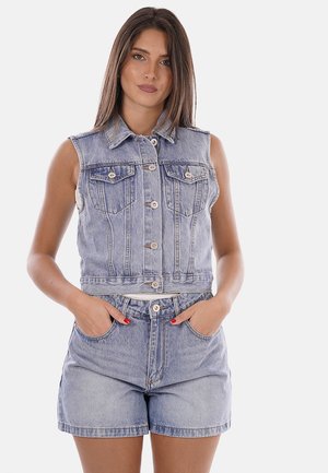 Mujer joven con cabello lacio que lleva un chaleco vaquero sin mangas con botones y shorts vaqueros de talle alto con las manos en los bolsillos.