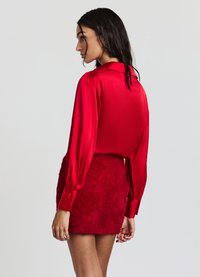 Blusa rossa di satin a maniche lunghe con colletto e spalle a sbuffo, abbinata a una mini gonna rossa testurizzata con motivi di rose.