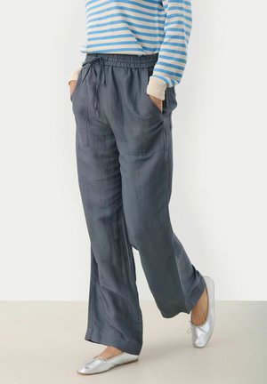 Persona con pantalones holgados de color gris oscuro con cordón, camiseta de manga larga a rayas azules y blancas, y bailarinas plateadas, con las manos en los bolsillos, de pie sobre un suelo claro.