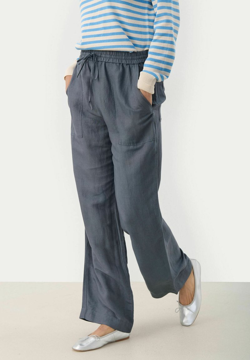 Personne portant un pantalon ample gris foncé à cordon, un haut à manches longues rayé bleu et blanc, et des ballerines argentées, les mains dans les poches, debout sur un sol clair.