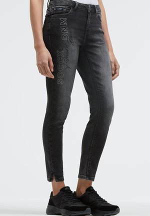 Slim fit black washed jeans with side text "SOCCX DNM" paired with black sneakers, shown from mid-thigh to feet.