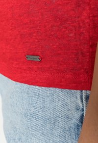 Top in tessuto a maglia rosso con texture, caratterizzato da un'etichetta metallica "Pepe Jeans" sull'orlo, abbinato a jeans in denim azzurro chiaro.