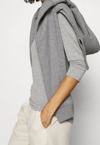 Camicia lunga grigia in cotone con maniche lunghe, sovrapposta a un leggero hoodie abbinato, indossata con pantaloni chiari; texture liscia e vestibilità comoda.