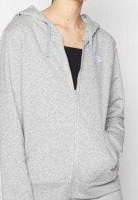Šedá mikina na zip z měkkého materiálu, s přední kapsou, nastavitelnými šňůrkami na kapuci a malým logem na hrudi.