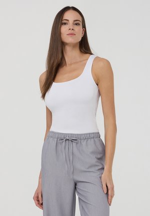 Donna con lunghi capelli castani che indossa un top bianco senza maniche e pantaloni grigio chiaro con coulisse, in piedi davanti a uno sfondo chiaro uniforme.