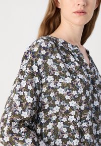 Blusa floreale con sfondo blu navy, caratterizzata da motivi di fiori bianchi e verdi, con scollatura bottonata e tessuto leggero.