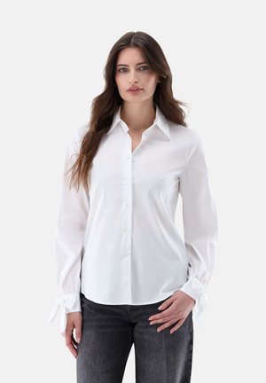Frau mit langen braunen Haaren, die eine weiße Bluse mit Knöpfen und gebundenen Manschetten sowie dunkelgraue Jeans trägt und vor einem schlichten hellen Hintergrund steht.