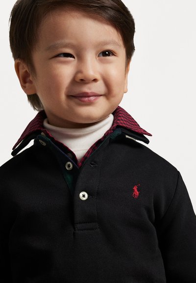 Μαύρο πόλο μπλουζάκι με κόκκινο κεντημένο λογότυπο, επάνω από ένα λευκό πουλόβερ με ψηλό λαιμό. Ο γιακάς είναι καρό, με κόκκινες και πράσινες αποχρώσεις.