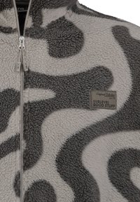 Fleece-Reißverschlussjacke in Grau und Creme mit abstraktem Muster, hohem Kragen und einem Markenlabel auf der linken Seite. Weicher, strukturierter Stoff.