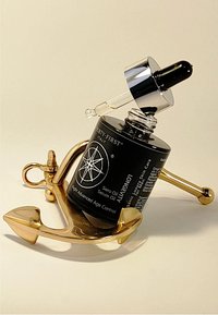 Zwart serum olie fles met een zilveren druppelaar, rustend op een gouden anker-vormige standaard, met tekst- en logo-details.