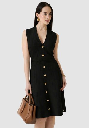 Donna che indossa un vestito nero senza maniche con bottoni, tiene una borsa marrone, con orecchini e bracciale d'oro su uno sfondo chiaro e semplice.