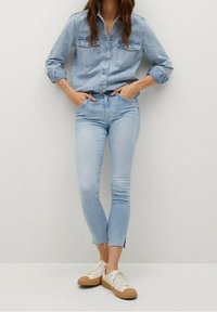 Mujer vestida con una camisa de mezclilla azul claro y jeans con dobladillos cortos, con las manos en los bolsillos, de pie contra un fondo claro y liso, con zapatillas blancas.