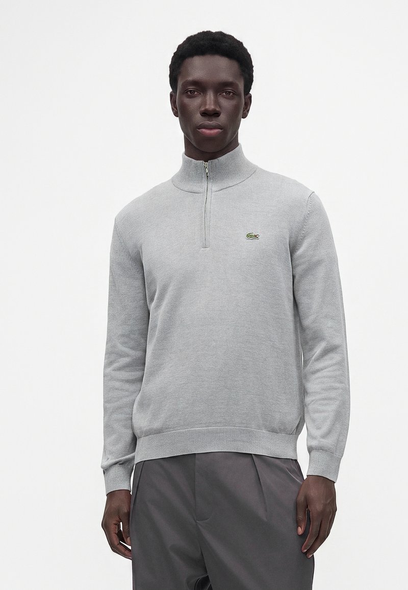 Homme portant un pull gris clair à fermeture éclair quart avec un petit logo crocodile vert et un pantalon gris foncé, debout devant un fond blanc.