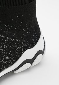 Baskets style chaussettes tricotées noires avec détail de semelle texturée blanche et noire sur fond blanc.