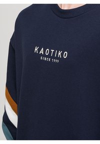 Felpa blu navy con colletto a girocollo a coste, con ricami "KAOTIKO" e "SINE 1999". Accenti a righe multicolori sulle maniche.