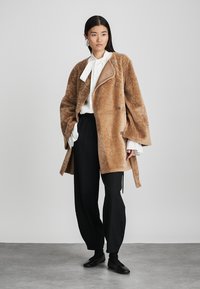 Brown faux fur coat with oversized sleeves, front buttons, and a contrasting brown collar, worn over a white blouse and black trousers.
