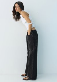 Frau mit langen dunklen Haaren lehnt sich nach vorne und trägt ein weißes ärmelloses Oberteil, schwarze Jeans mit weitem Bein und schwarze Sandalen vor einer einfarbigen Wand.