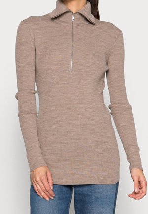 Pull à manches longues beige côtelé avec col à demi-fermeture éclair, porté avec un jean bleu, visible de la nuque aux hanches, bras détendus sur les côtés.