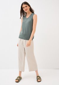 Une femme se tient avec confiance, portant un haut sans manches vert, un pantalon beige et des sandales vertes, avec un fond neutre mettant en valeur sa tenue.