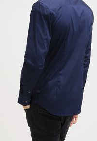 Man som bär en figurnära marinblå långärmad skjorta och svarta jeans, står med en hand i fickan och vänder ryggen mot kameran.