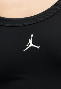 Κοντινό πλάνο μαύρης λαιμόκοψης πουκάμισου με λευκό κεντητό λογότυπο Jumpman, που δείχνει έναν μπασκετμπολίστα στον αέρα να κρατά μια μπάλα.