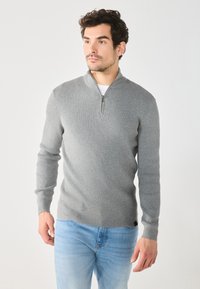Sweater gris con cuello de cremallera y textura acanalada, de mangas largas y diseño ajustado. Combinado con jeans azul claro. Sutil marca en el dobladillo.