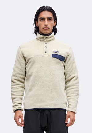 Uomo con capelli scuri che indossa un pullover in pile beige con colletto alto e tasca sul petto blu navy con bottoni, in piedi davanti a uno sfondo chiaro e uniforme.