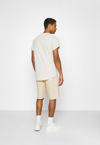Beige kortermet t-skjorte med rullet ermer, sammen med lysebeige shorts. Hvite joggesko med subtil merkedesign. Nøytral bakgrunn.