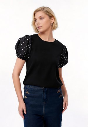 Femme blonde portant un haut noir avec des manches bouffantes noires à pois blancs et un jean bleu foncé taille haute avec des boutons décoratifs.