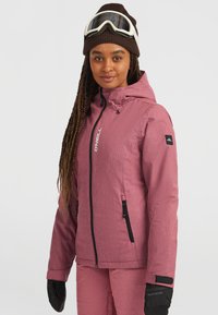 Vrouw in een roze O'Neill skijas en -broek, bruine beanie met witte skibril en zwarte handschoenen, kijkt recht vooruit.