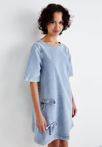 Svetlo modra denim obleka s kratkimi rokavi, okroglim izrezom in dvema sprednjima žepoma z nagubanim detajlom. Mehka tekstura in ohlapen kroj.