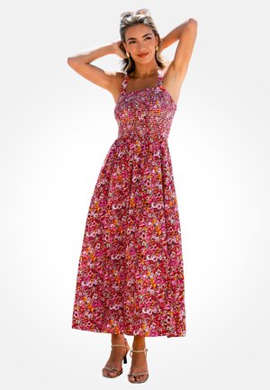 Woman wearing a sleeveless, pink floral maxi dress and heels, posing with hands behind her head and sunglasses on top of her hair.