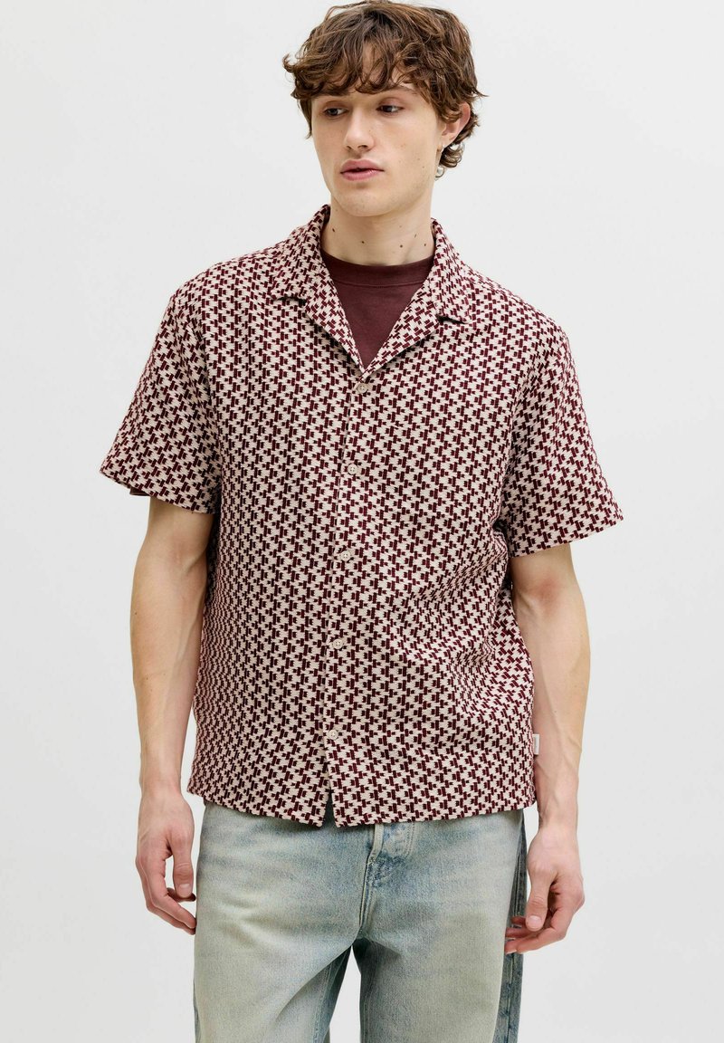 Jeune homme portant une chemise à manches courtes boutonnée bordeaux à motifs, par-dessus un t-shirt bordeaux foncé, avec un jean bleu clair.