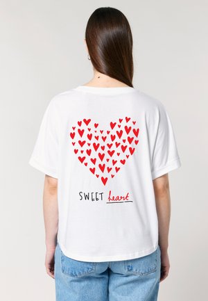 Auf dem Bild ist der Rücken einer Person zu sehen, die ein weißes T-Shirt mit einem Herzdesign aus roten Herzen trägt. Darunter steht der Text "SWEET heart".