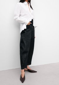 Valkoinen nappipaitapusero, mustat korkeavyötäröiset leveälahkeiset farkut ja ruskeat kärjelliset mokkasiinit vaaleaa taustaa vasten.