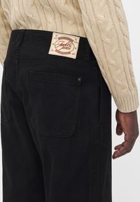 Personne portant un jean en denim noir avec une poche arrière et un pull beige en maille torsadée, affichant une étiquette de la marque Jack & Jones sur la ceinture.