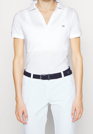 Frau trägt weißes Poloshirt mit kurzen Ärmeln und "CK"-Logo auf der Brust, weiße Hose und einen dunklen gewebten Gürtel mit silberner Schnalle.