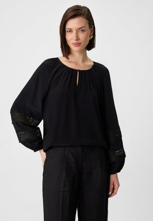 Blusa nera con scollatura a chiave e accenti in pizzo nero testurizzato sulle maniche. Tessuto morbido che drappeggia, abbinata a pantaloni scuri.