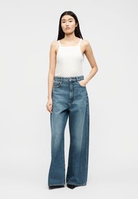 Νεαρή γυναίκα με μακριά σκούρα μαλλιά, ντυμένη με λευκό τοπ και φαρδιά μπλε τζιν παντελόνια, στέκεται με το ένα χέρι στην τσέπη.