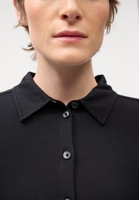 Černá košile s límečkem a hladkou texturou, která má zapínání na knoflíky a černé knoflíky. Detailní pohled na límec a výstřih.