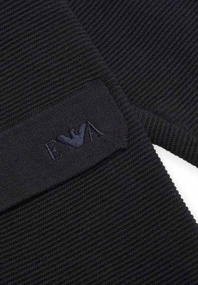 Tessuto a coste nero con una superficie texturizzata, caratterizzato da un patch rettangolare con ricamo "EVA" in un colore scuro e un design stilizzato di un uccello.